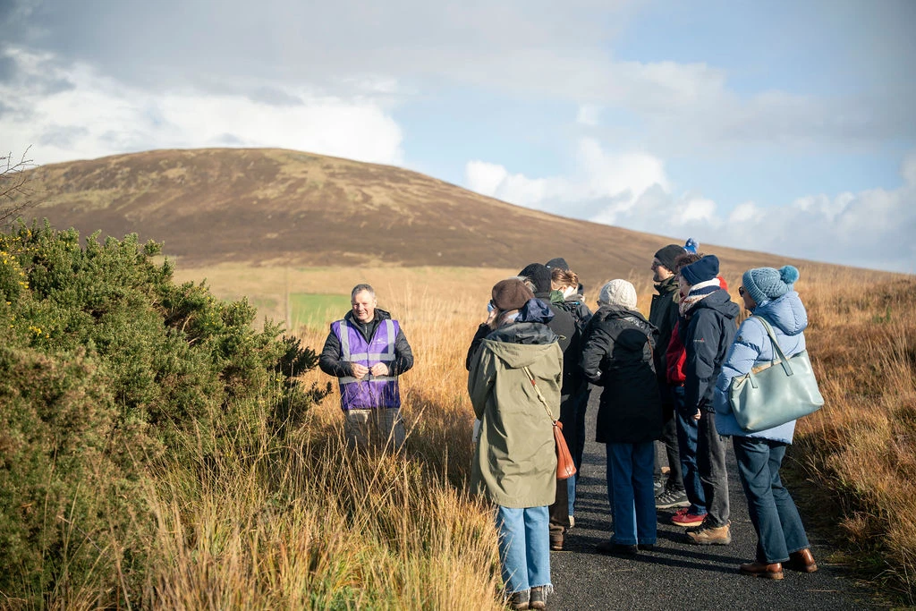 Group on guided nature walk in countryside, learning about local plants, with hills in the background.