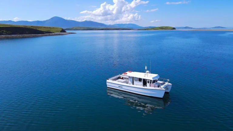 Boat cruising clear waters against scenic Irish landscape at Mayo Dark Sky Festival, under bright blue sky.