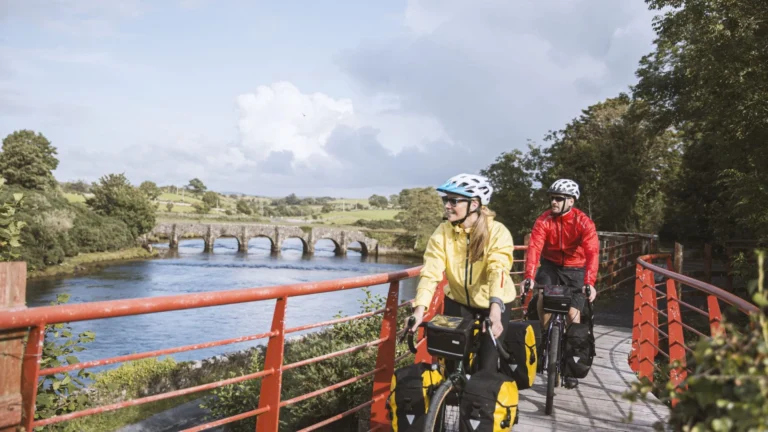 Cyclists on scenic riverside path in County Mayo, Ireland, with picturesque stone bridge in view.