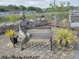 Statue of a woman on a bench by the river, surrounded by plants, at Europe’s Mayo Dark Sky Festival site.